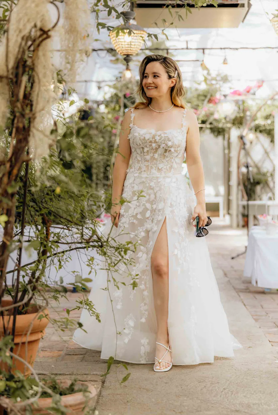 Eine Braut lächelt, während ich sie als Hochzeitsfotograf München bei ihrem Gang durch die helle Gärtnerei fotografiere.
