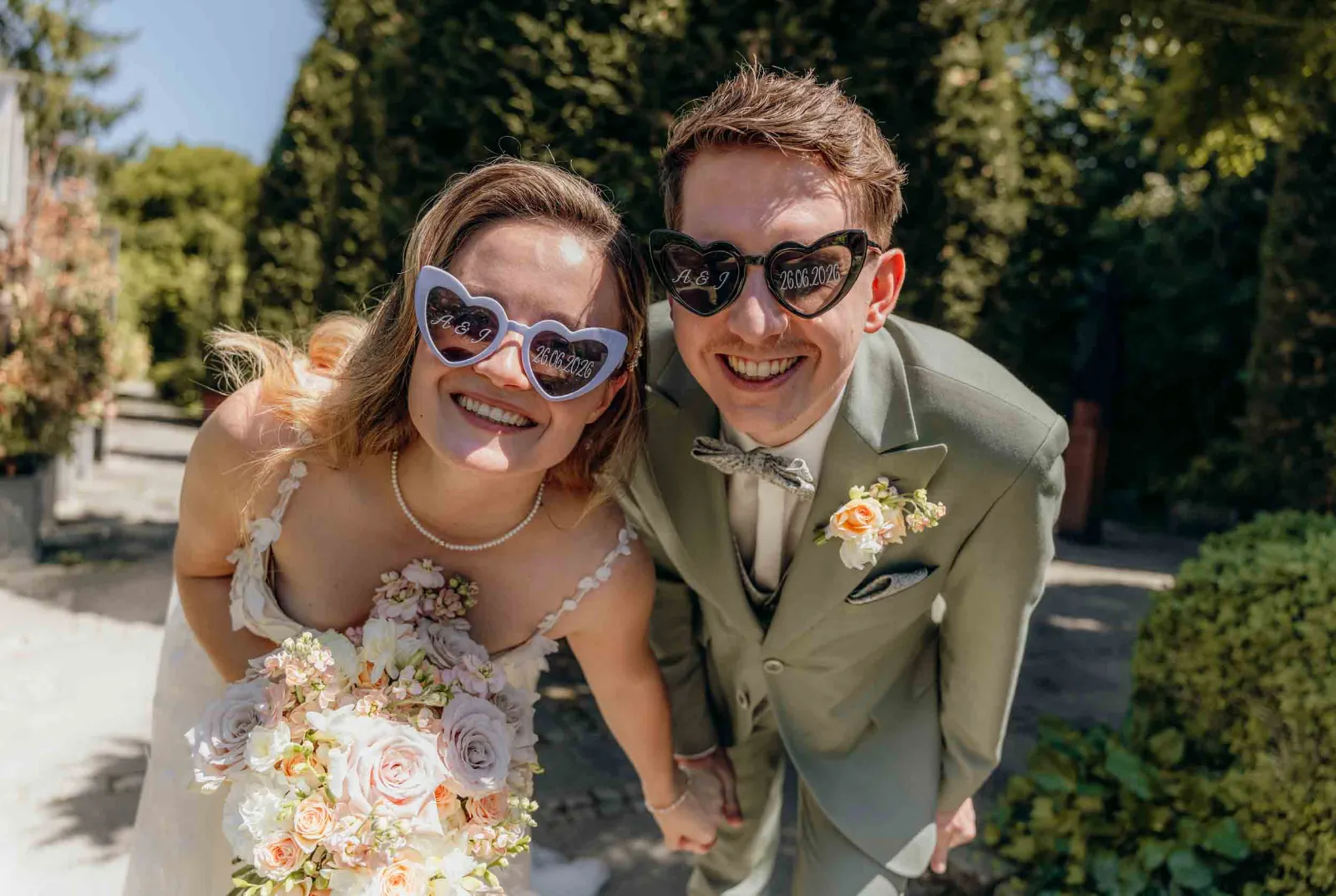 Ein lachendes Brautpaar trägt bei seiner Hochzeit als Hochzeitsfotograf München MOKATI herzförmige Sonnenbrillen vor grüner Kulisse in der alten Gärtnerei in Taufkirchen bei München.