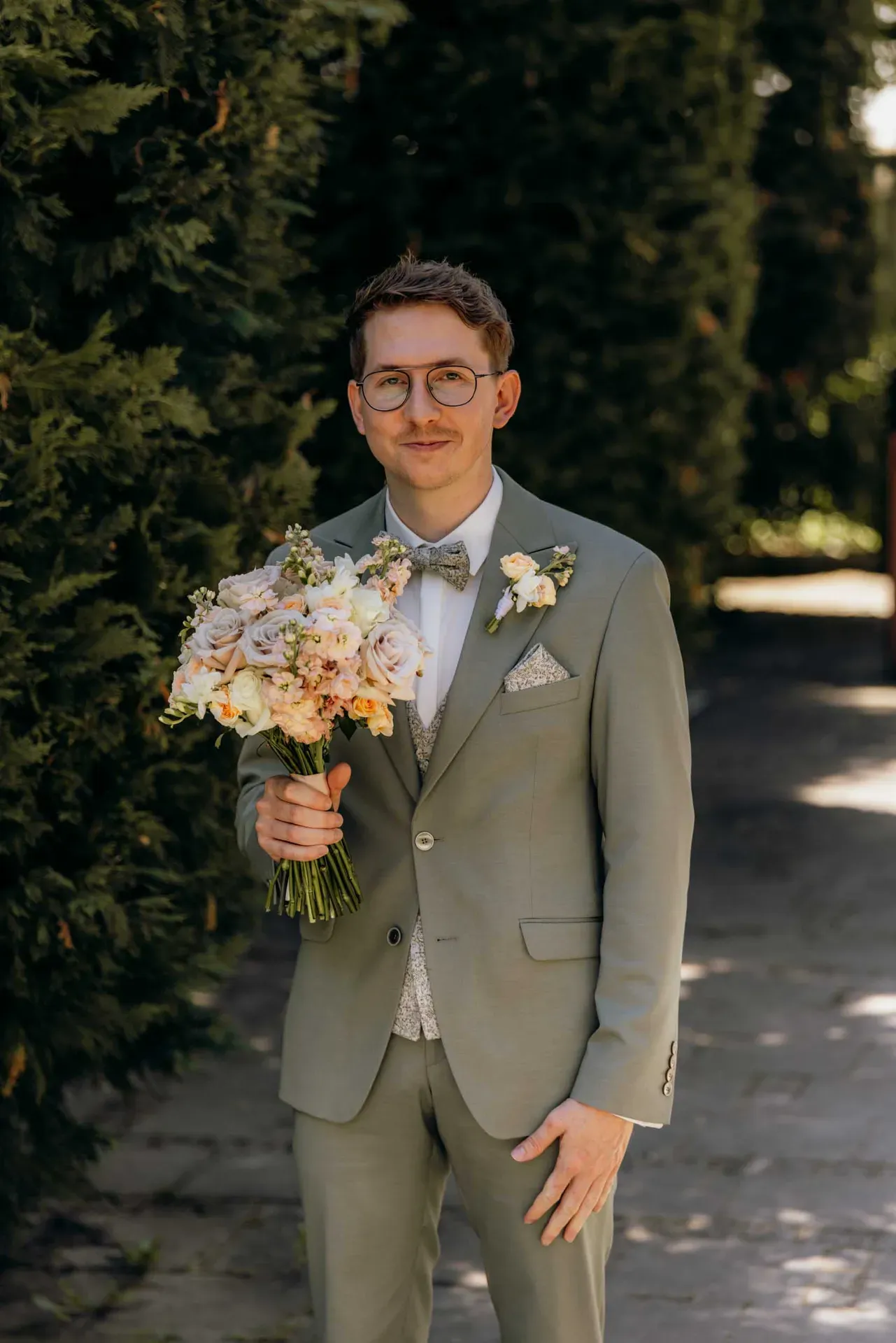 Ein Bräutigam im hellgrünen Anzug hält bei einer kleinen Hochzeit den Brautstrauß mit hellen Blüten in die Kamera.