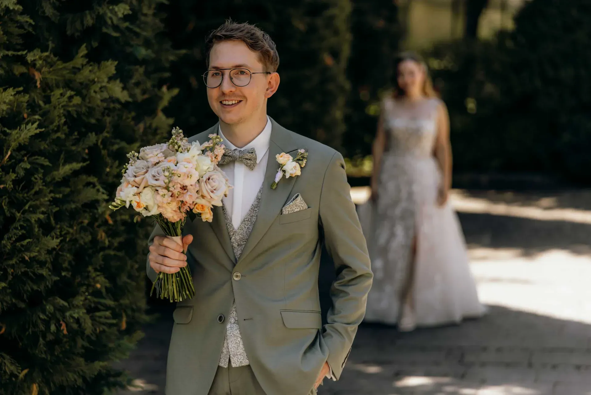 Ein Bräutigam hält im Hintergrund die Braut im Blick, während er den Blumenstrauß bei dieser kleinen Hochzeit in der Gärtnerei in den Fokus rückt.