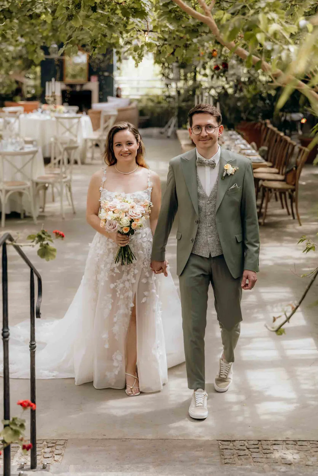 Ein Brautpaar geht Hand in Hand durch die gärtnerische Kulisse ihrer kleinen Hochzeit und lächelt freundlich in die Kamera.