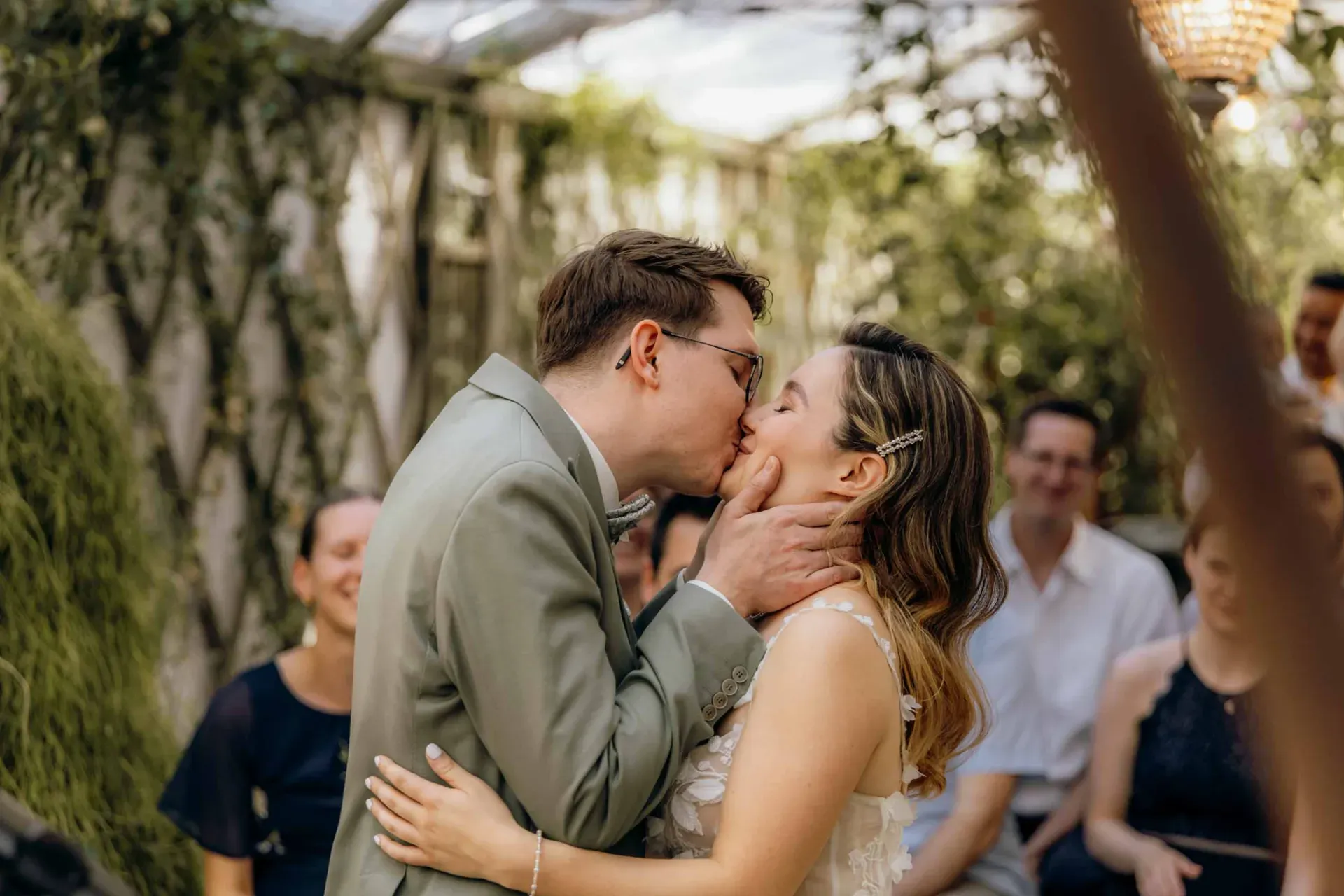 Ein Brautpaar tauscht während einer kleinen Hochzeit den ersten Kuss aus, während Gäste im Hintergrund aufmerksam zusehen.