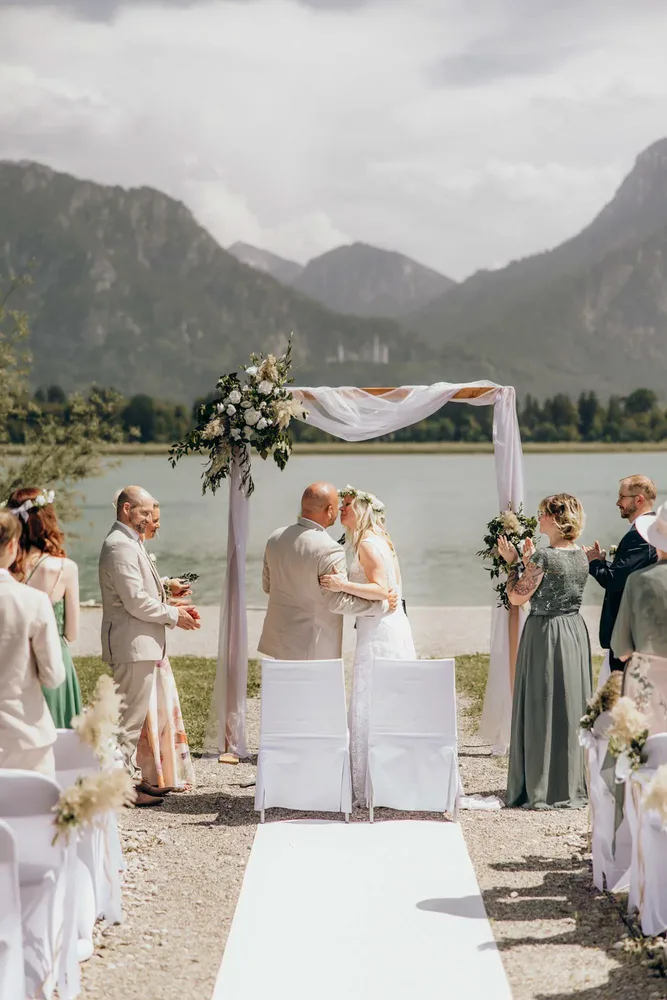 Brautpaar küsst sich unter einem Blumenbogen mit Bergen und See im Hintergrund. Hochzeitsfotograf München MOKATI