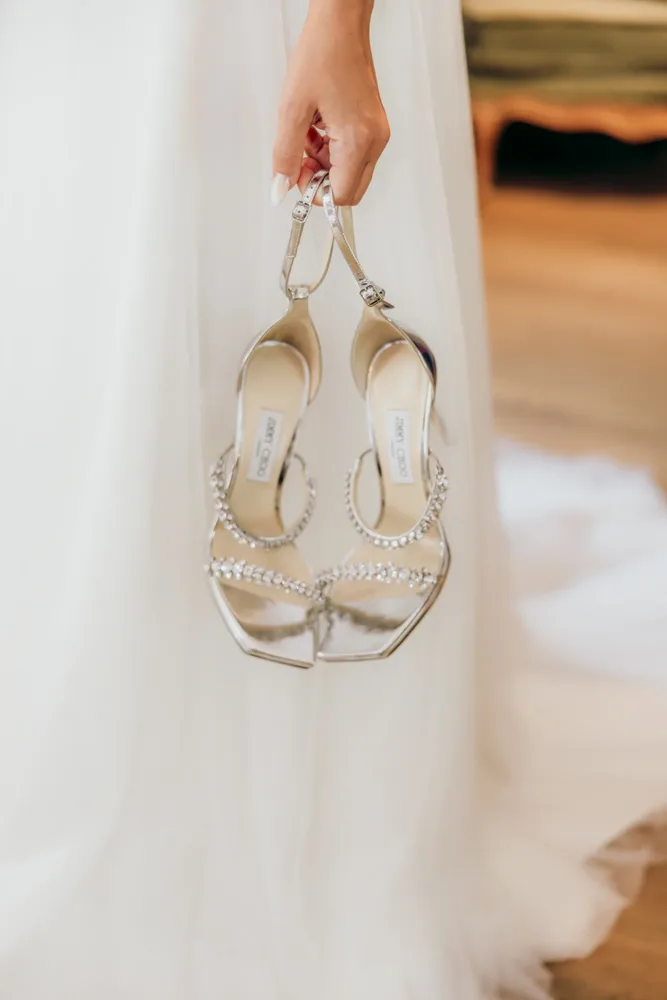 Eine Hand hält silberne Jimmy Choo Brautschuhe mit Strassbesatz. Hochzeitsfotograf MOKATI