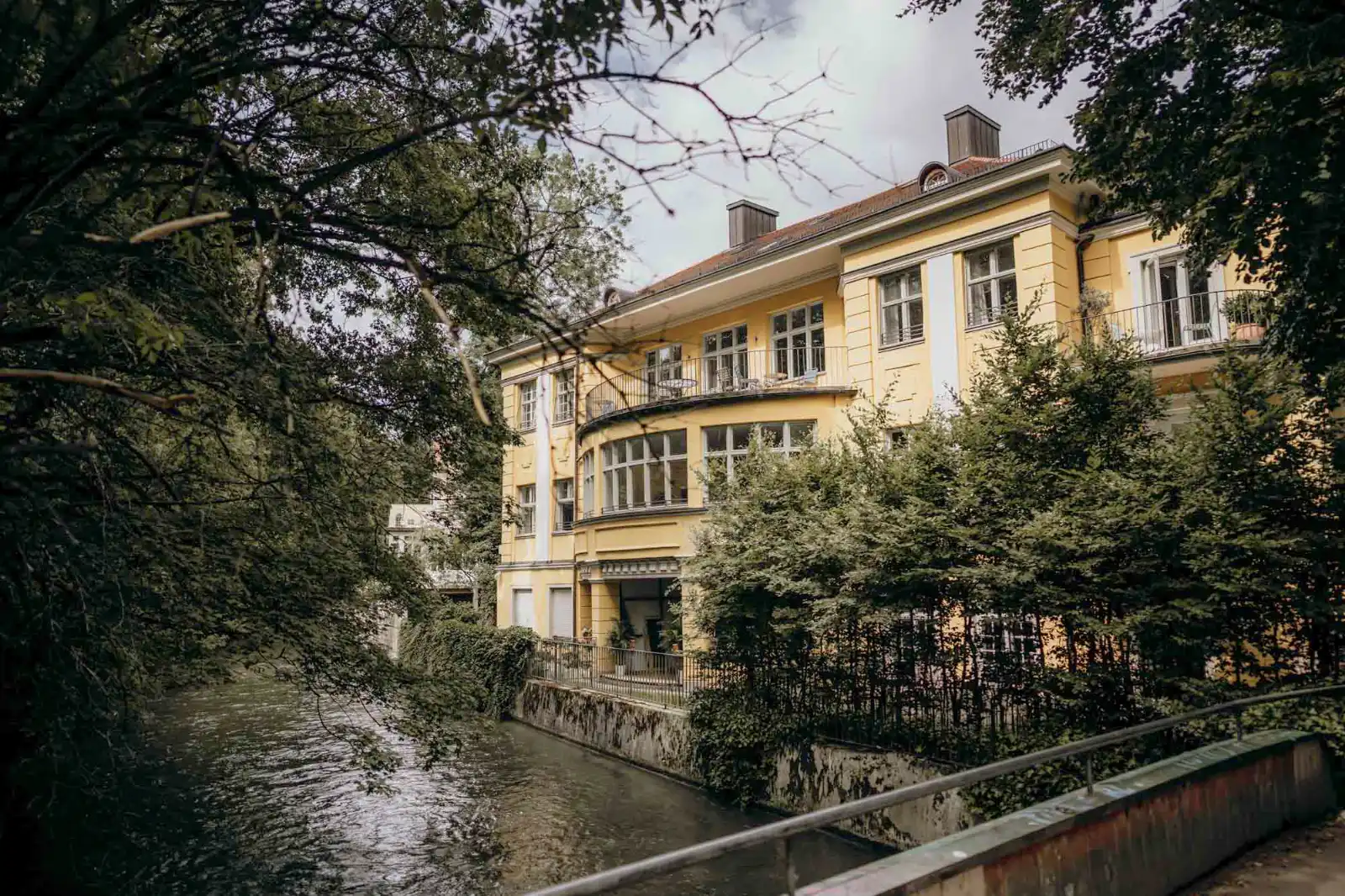 Das gelbe Gebäude des Standesamts Mandlstraße in München, davor der Bachlauf und alte Bäume