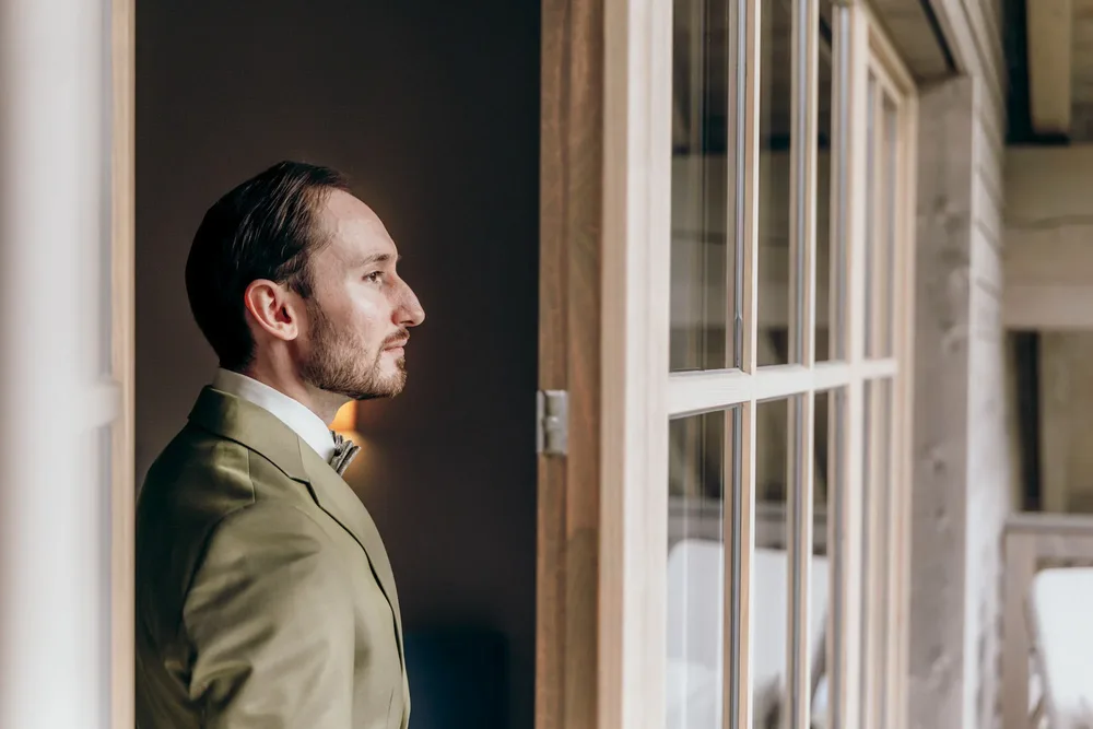 Mann im hellgrünen Anzug und Fliege blickt aus einem Fenster. Hochzeitsfotograf MOKATI