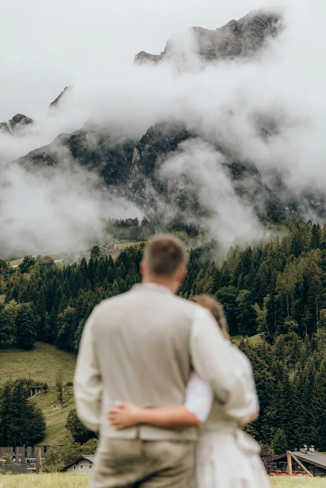 Paar von hinten, Arm in Arm, blickt auf bewölkte Berge und grüne Waldlandschaft. Hochzeitsfotograf MOKATI