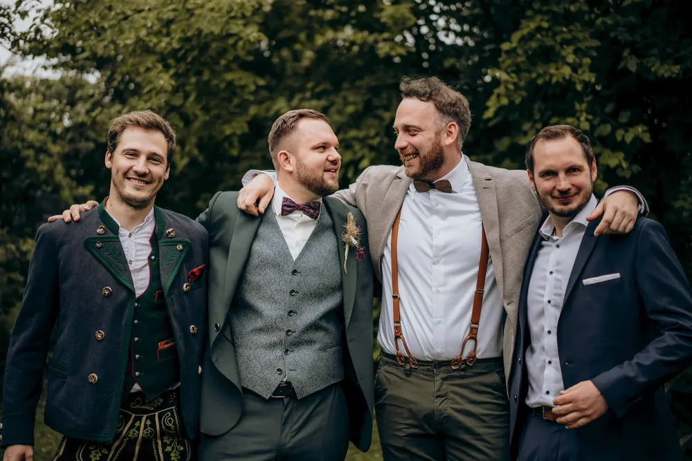 Vier Männer in Anzügen und Trachten stehen lächelnd, zwei umarmen sich. Hochzeitsfotograf MOKATI