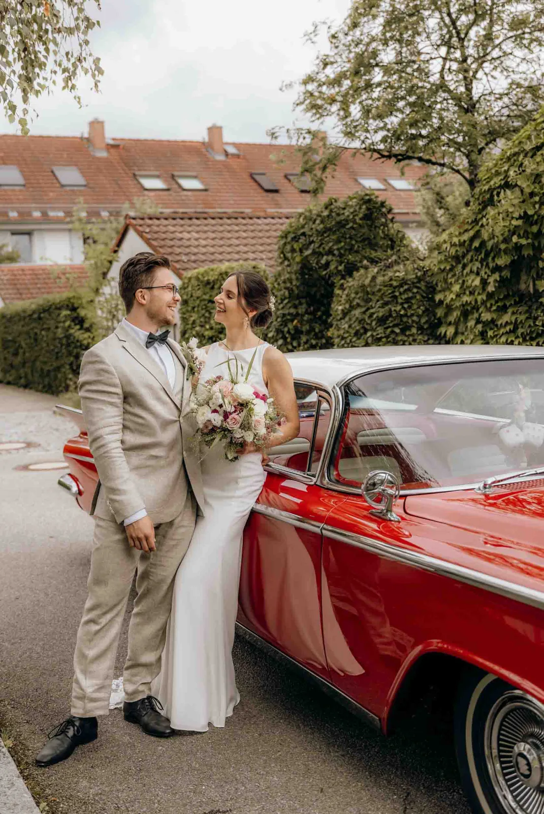 Ein Brautpaar lächelt sich an, während ich als Hochzeitsfotograf München diesen Moment an ihrem alten roten Auto in Poing festhalte.